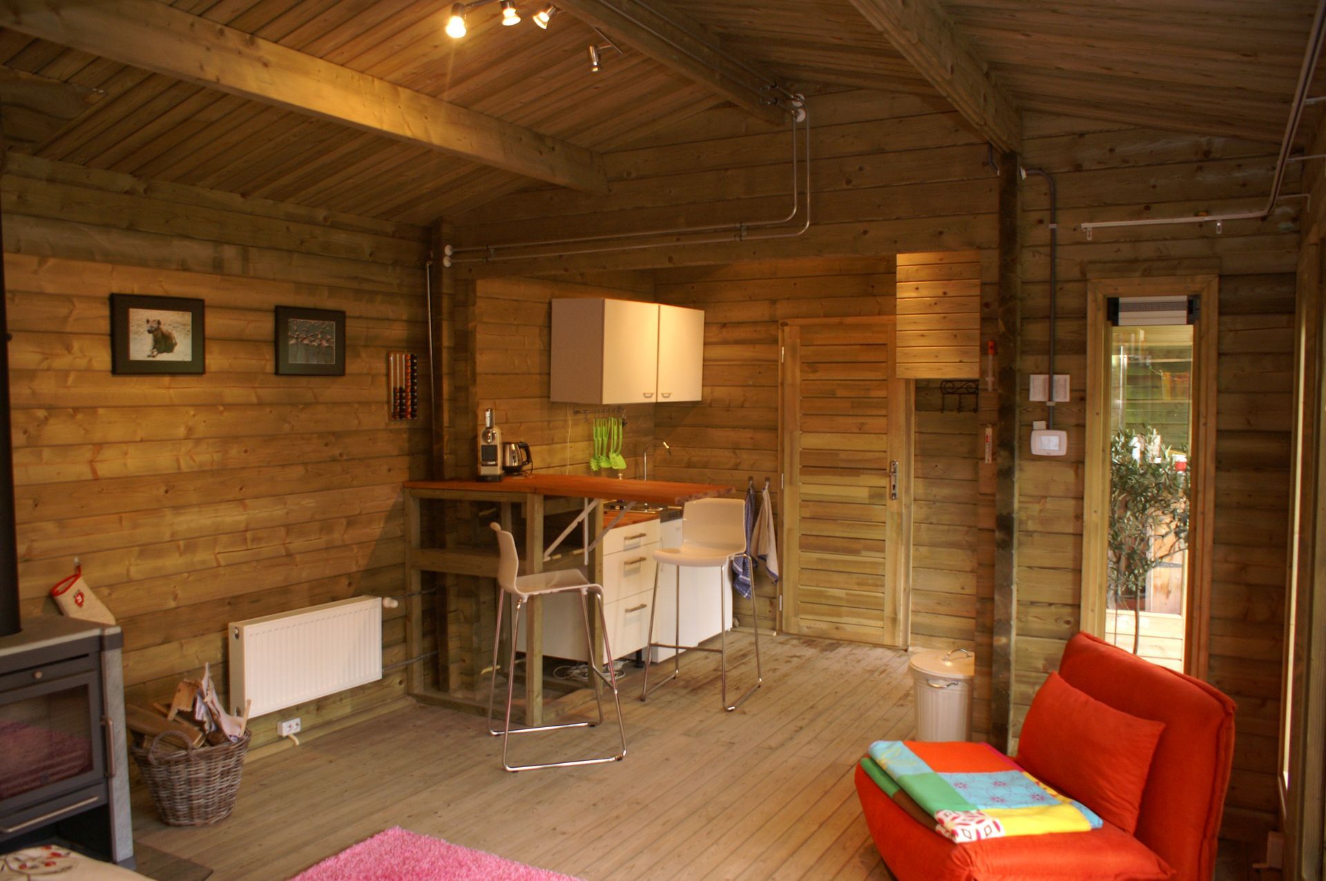 Houten hutinterieur met keukenhoekje, rode stoel en kleine tv bij een deur naar buiten.