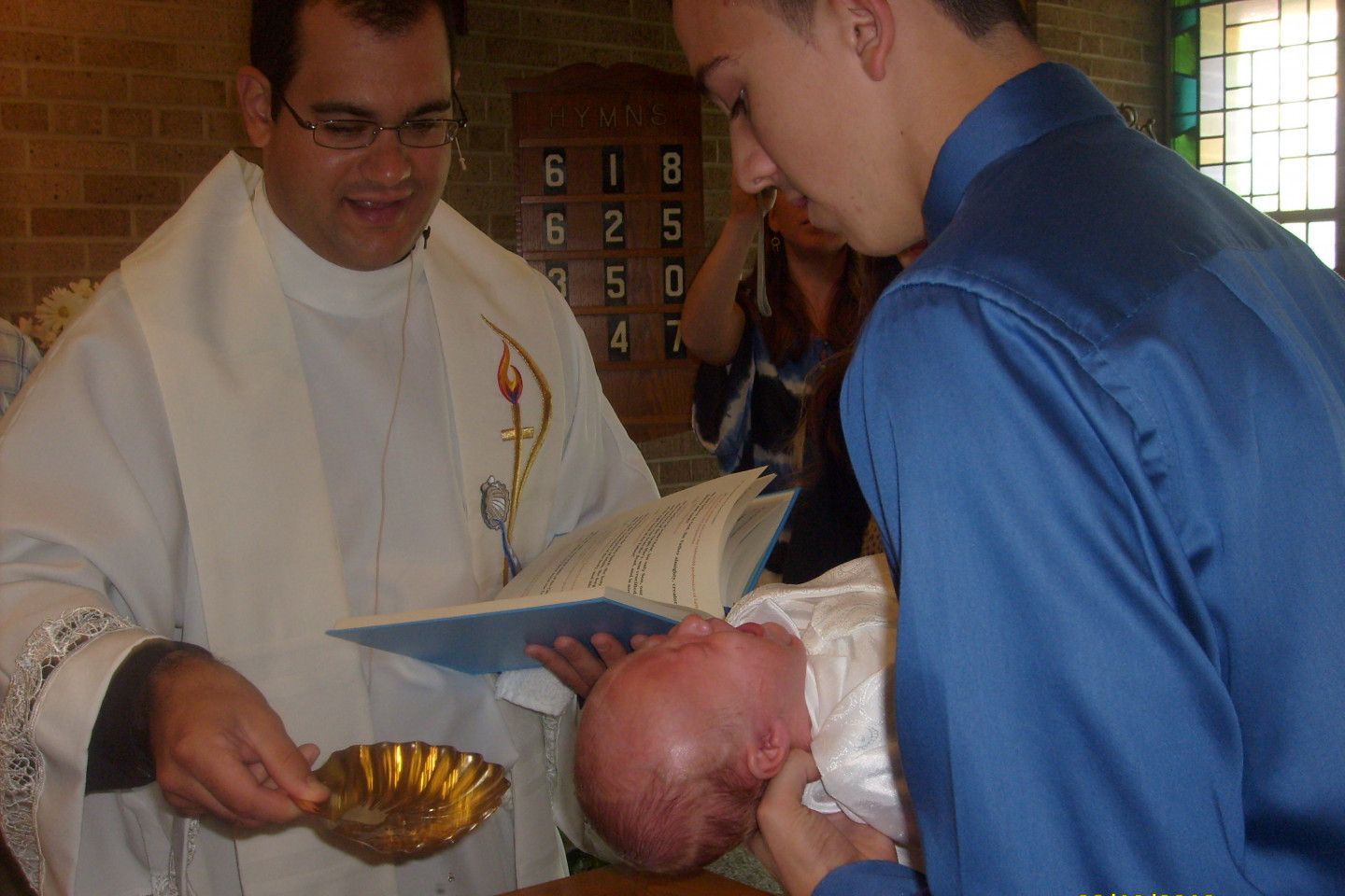 Priest baptizes a baby in a church, holding a golden bowl. Man in blue shirt assists.