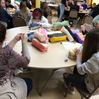 People gathered around a large round table, possibly crafting. Various fabric items are on the table.