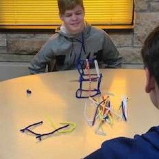 Two teens at table with pipe cleaner structures. One smiles, background yellow blinds.