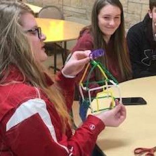Person in red jacket holding a small structure; two others watch at a table.