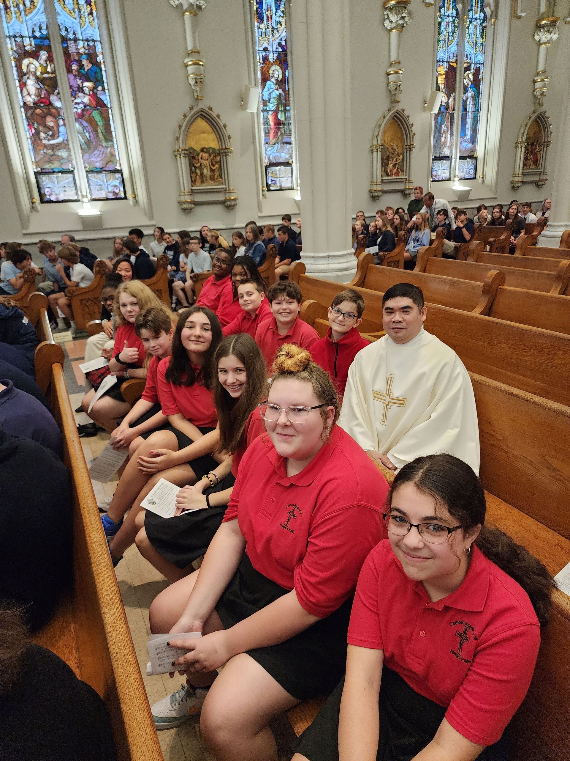 Group of people in red shirts sitting in pews in a church with stained glass windows. A priest is also present.