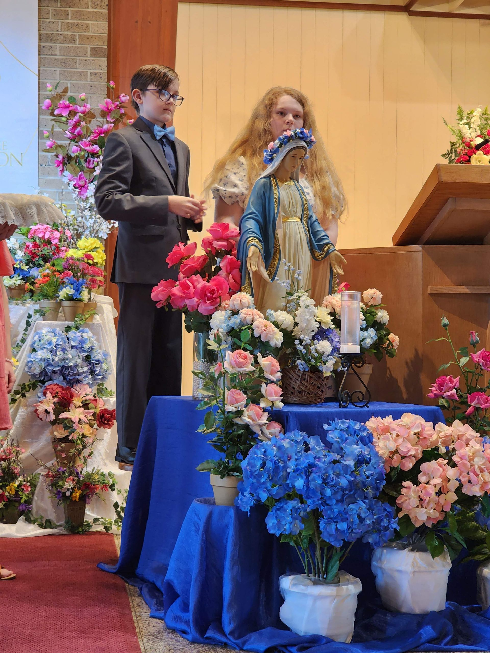 Boy and woman with statue, surrounded by flowers, in church setting.