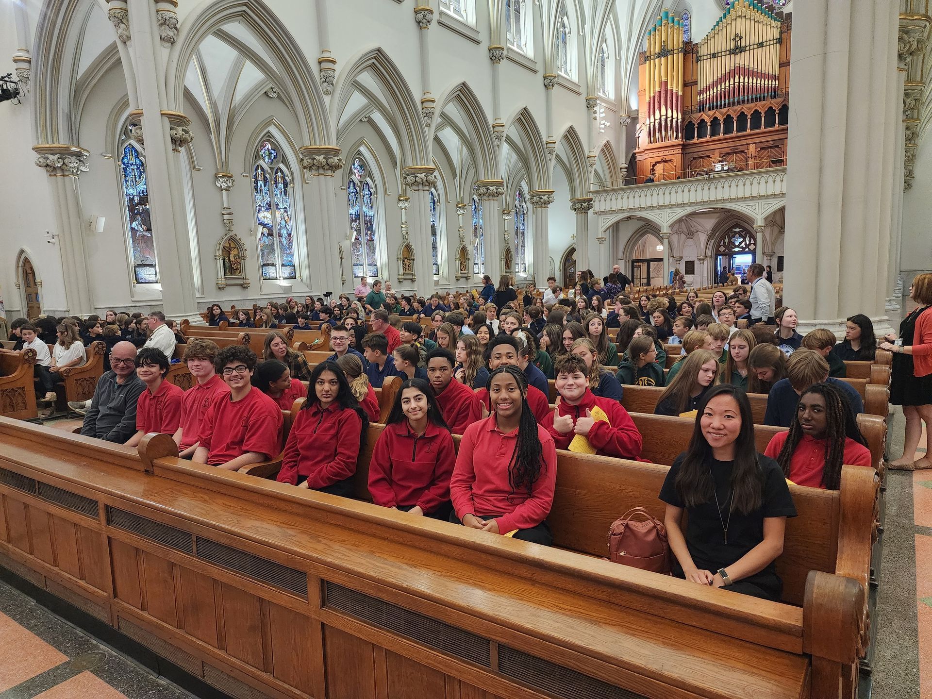 Students in red shirts seated in church pews during a service. Ornate architecture, stained glass windows.