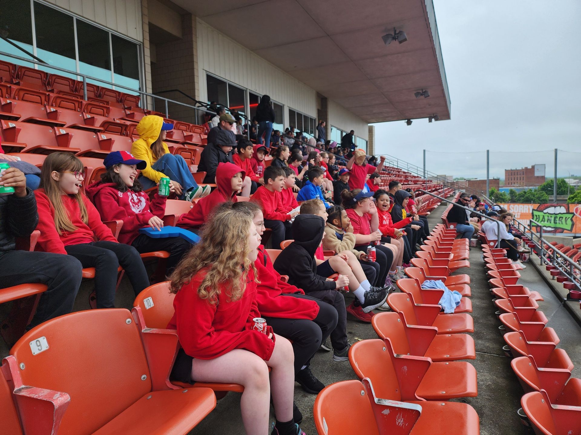 Children in red hoodies and jackets sit in stadium seats, watching an event.