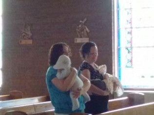 Two women in a church hold babies. One baby wears a lamb hat.
