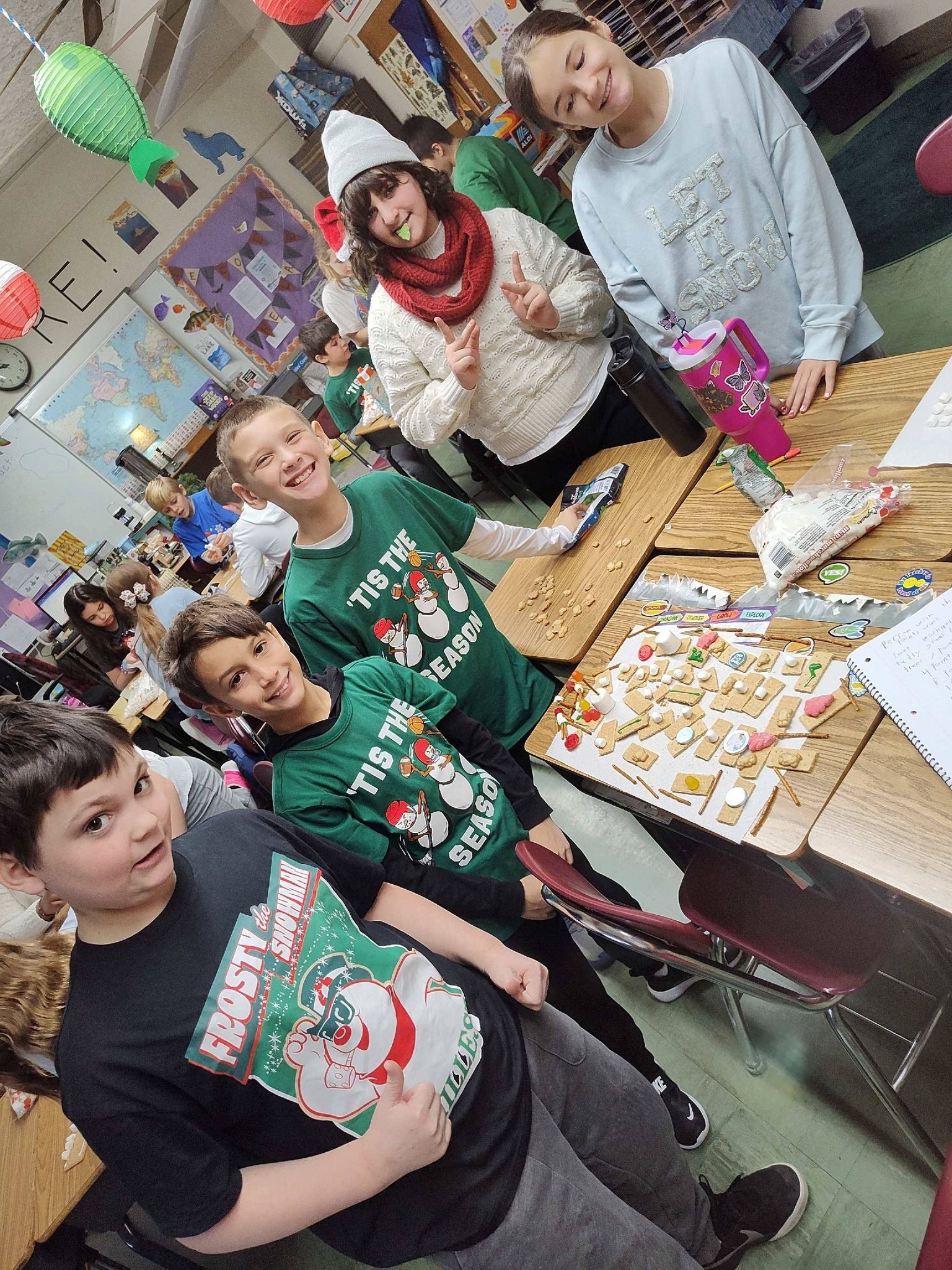 Children making treats at a classroom table, festive shirts, winter holiday decorations.