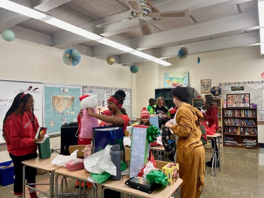 Classroom gift exchange with students wearing festive attire, desk covered in presents.