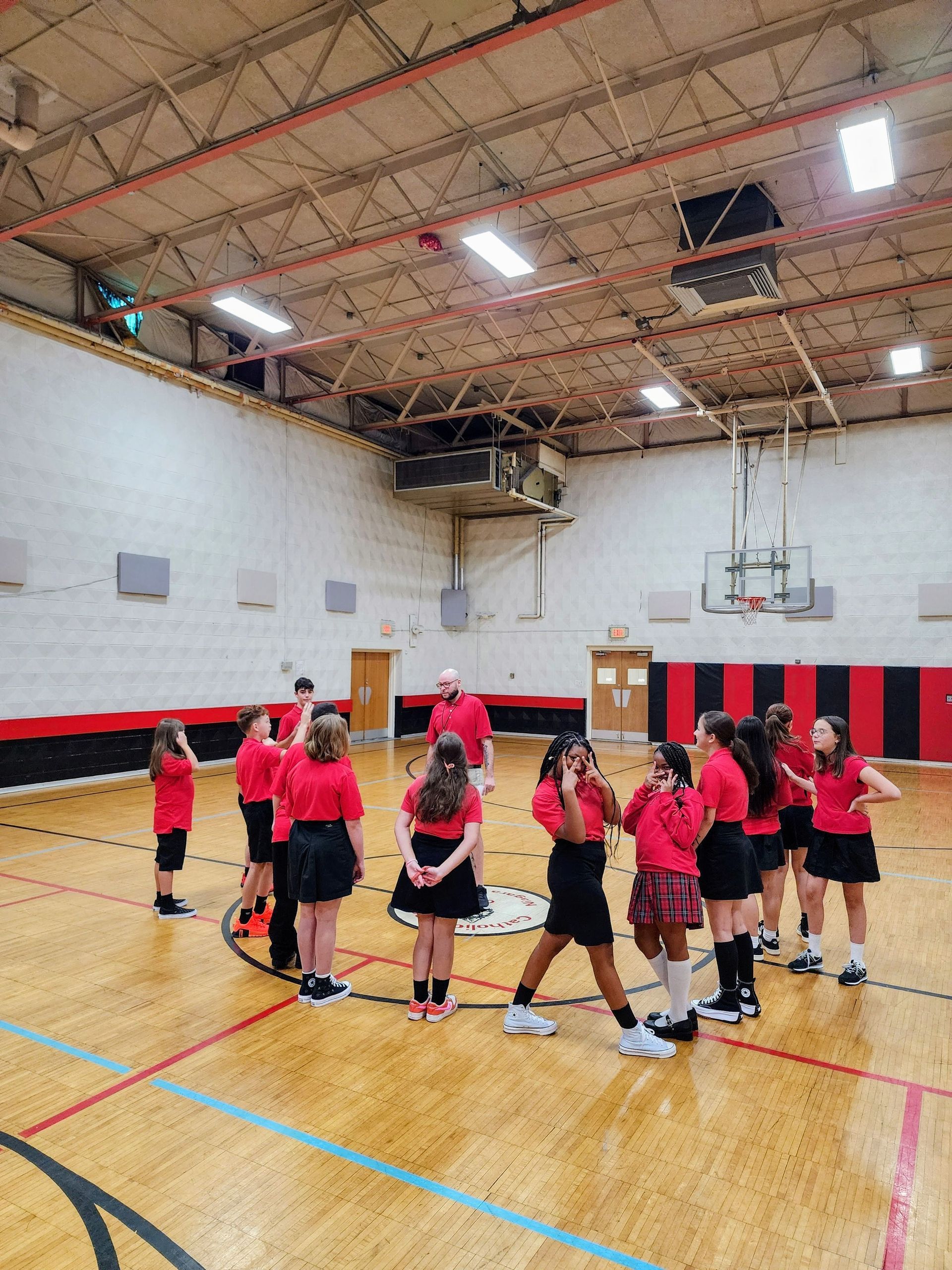 Students in red shirts and black skirts stand in a circle on a gym floor.