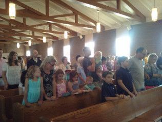 People stand in church pews, some holding children, with sunlight streaming through the windows.