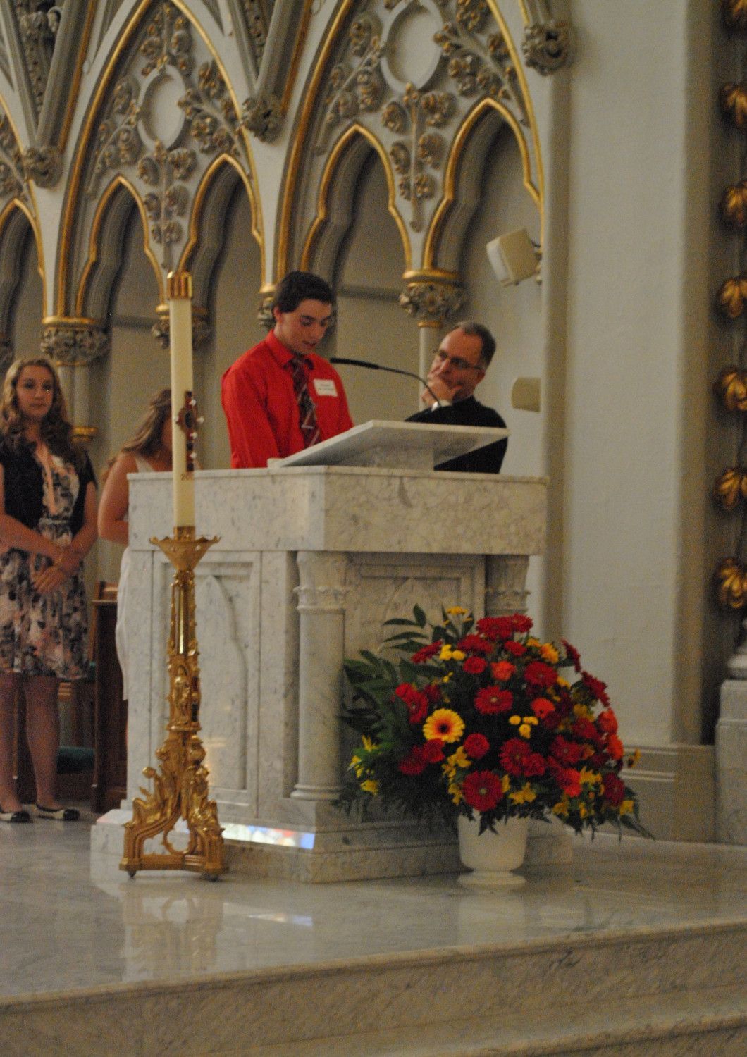 Man in red shirt reads at a podium with flowers in a church, another man smiles nearby.