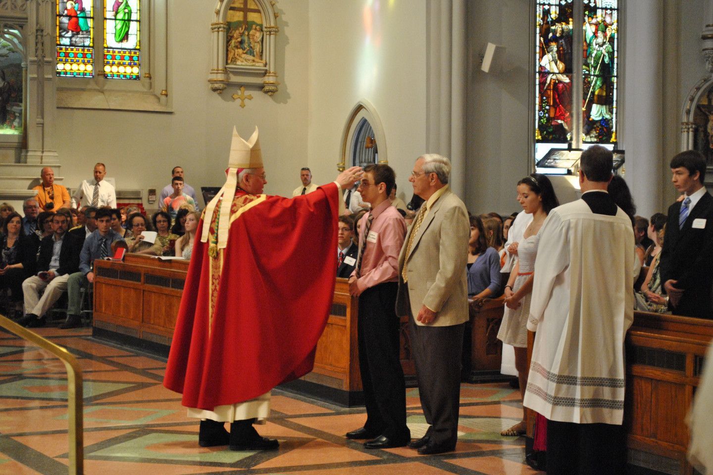 A bishop in red vestments anoints a person's forehead in a church ceremony.