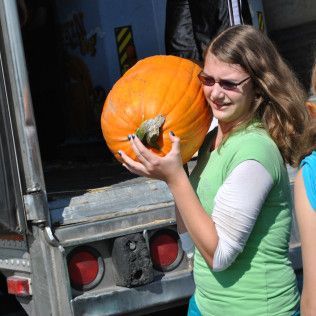 Woman carrying a large orange pumpkin at a pumpkin patch, by a truck.