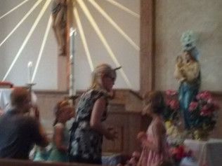People in a church, near a statue of Mary and baby Jesus.  A crucifix hangs in the background.