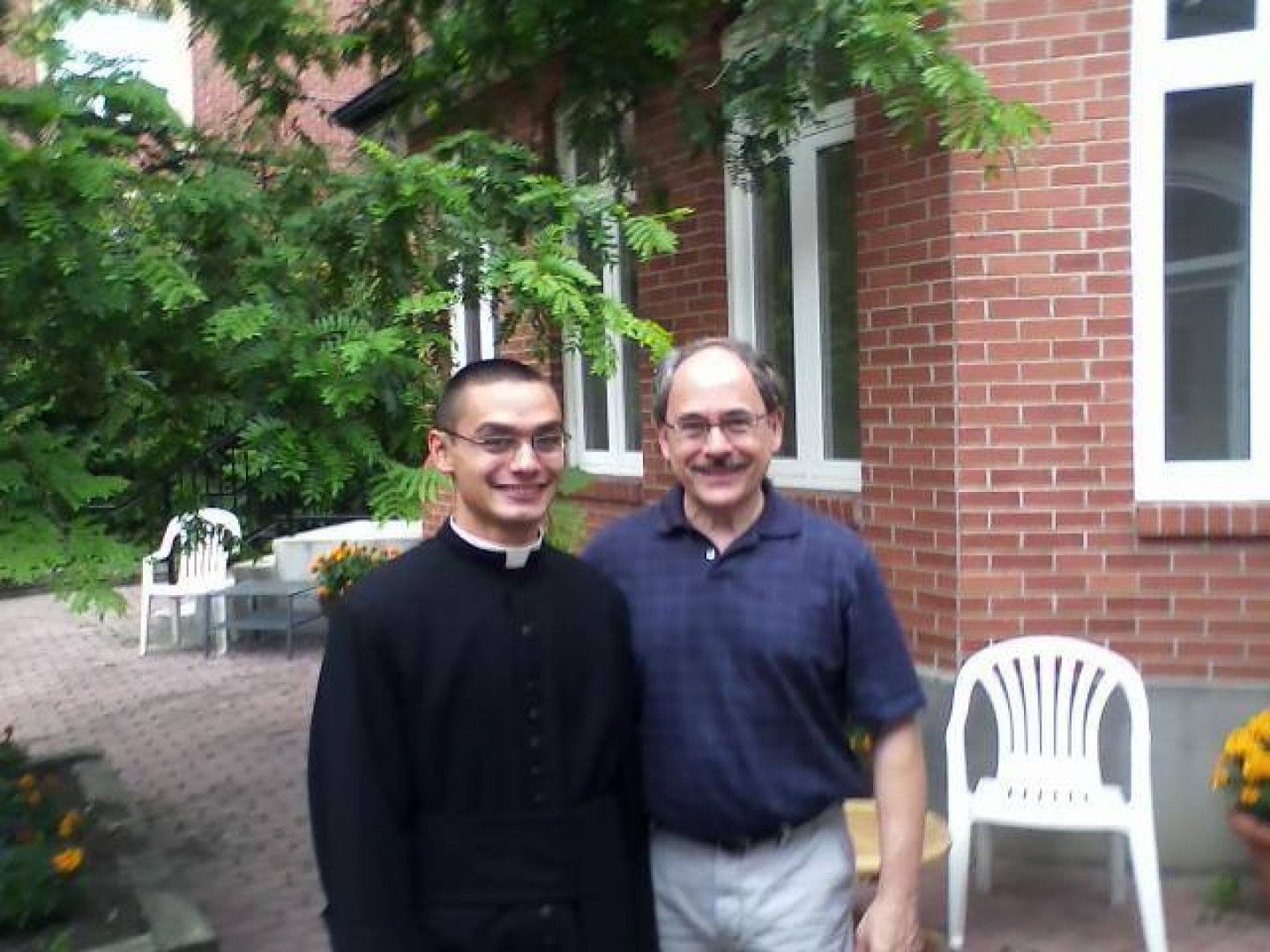 Two men smiling outdoors by a brick building. One wears a black clerical collar, the other a blue shirt.