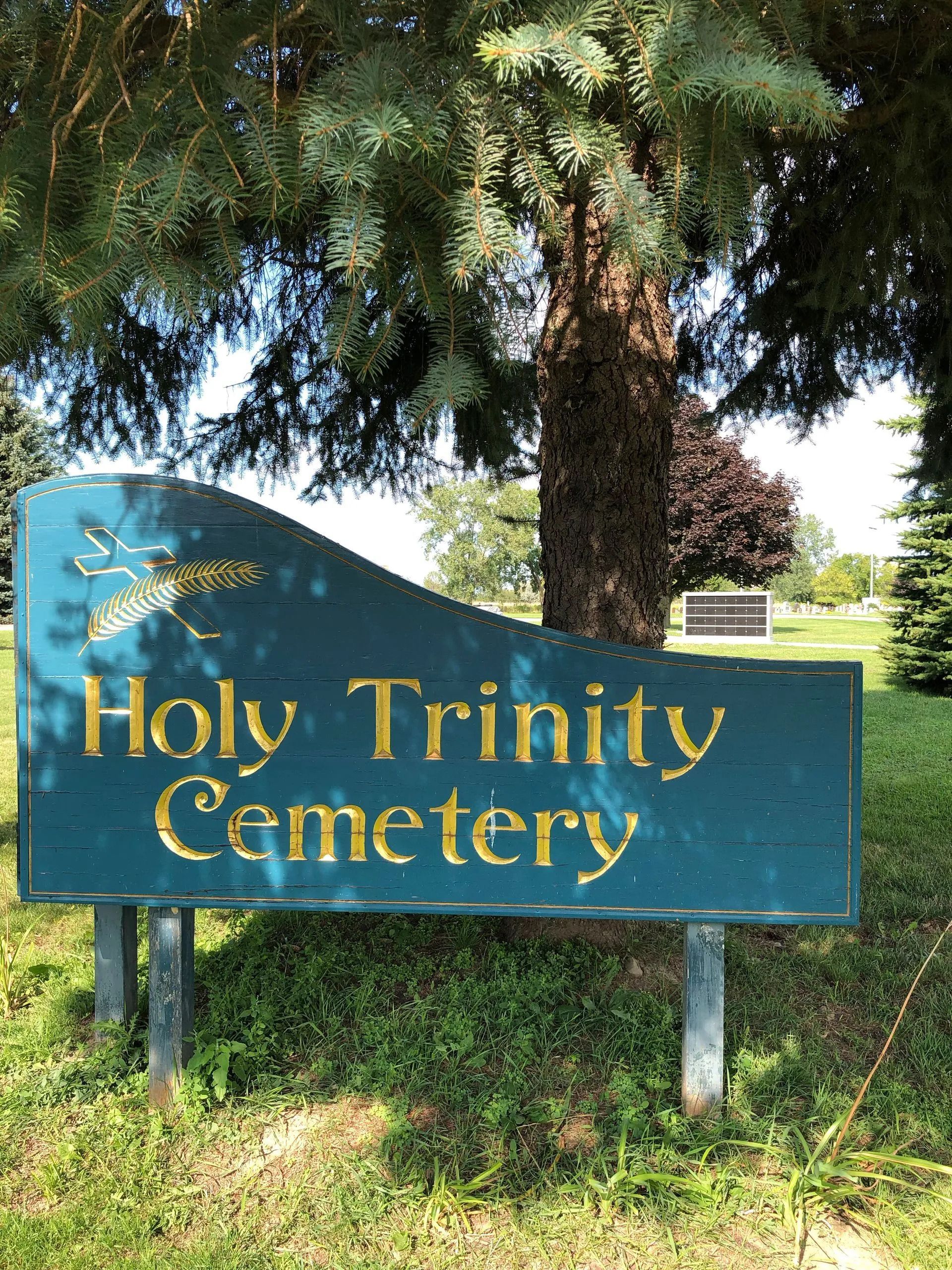 Sign for Holy Trinity Cemetery, teal with gold lettering. Green foliage surrounds.