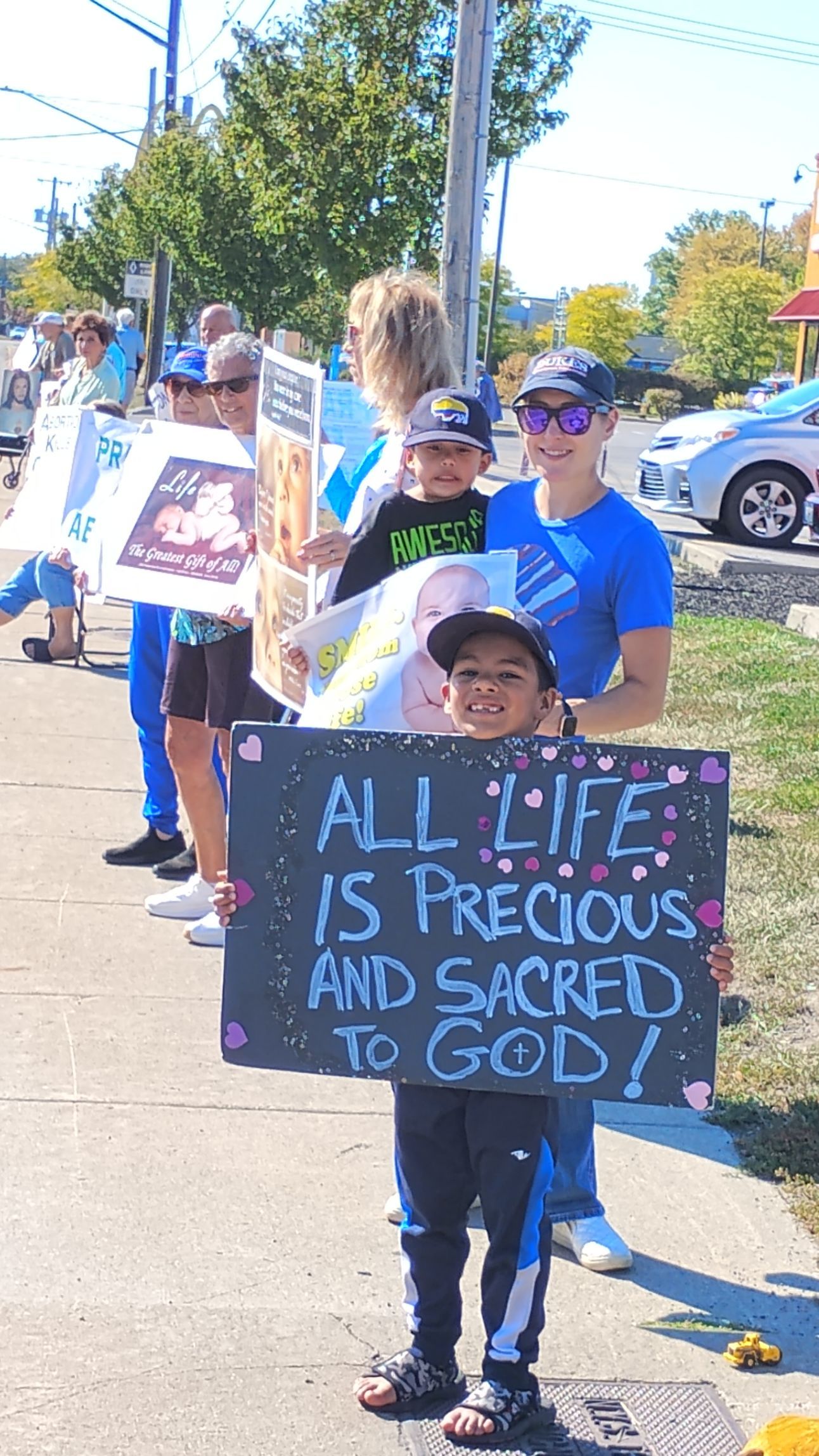People holding signs with messages, standing on a sidewalk. One child holds a sign reading 