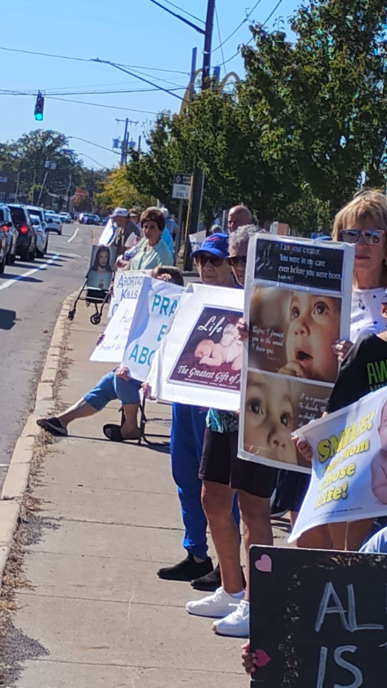 People on sidewalk holding signs with an anti-abortion message near a road with cars.