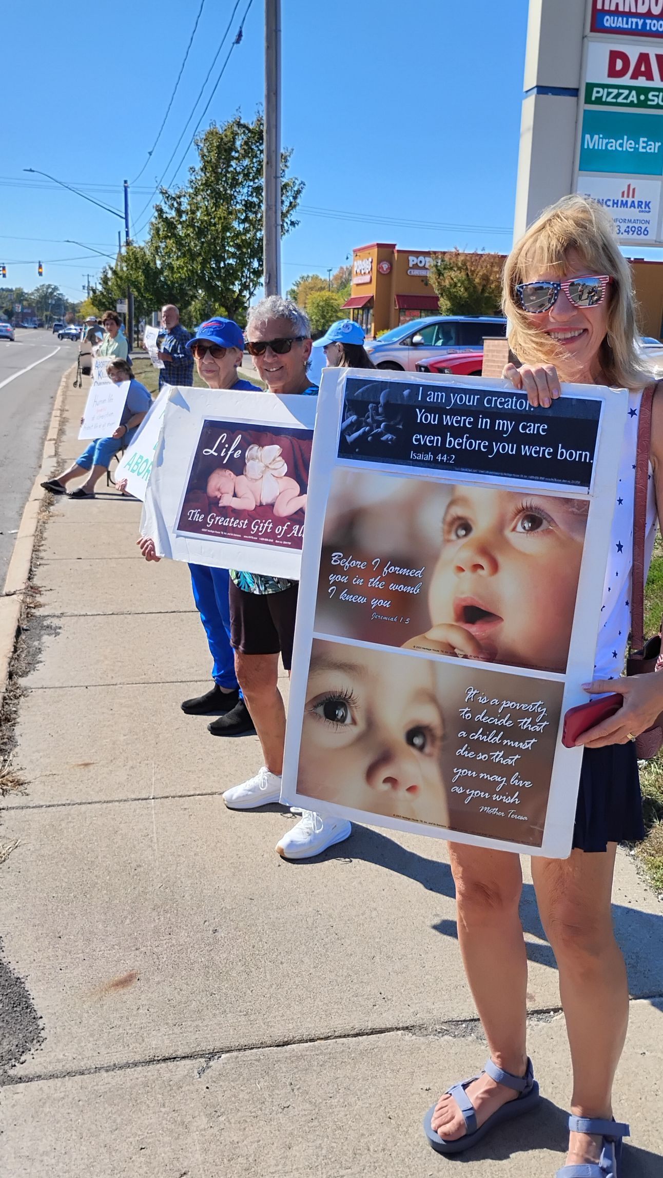 People holding signs with baby images, standing on sidewalk. One sign has text. Bright sunny day.