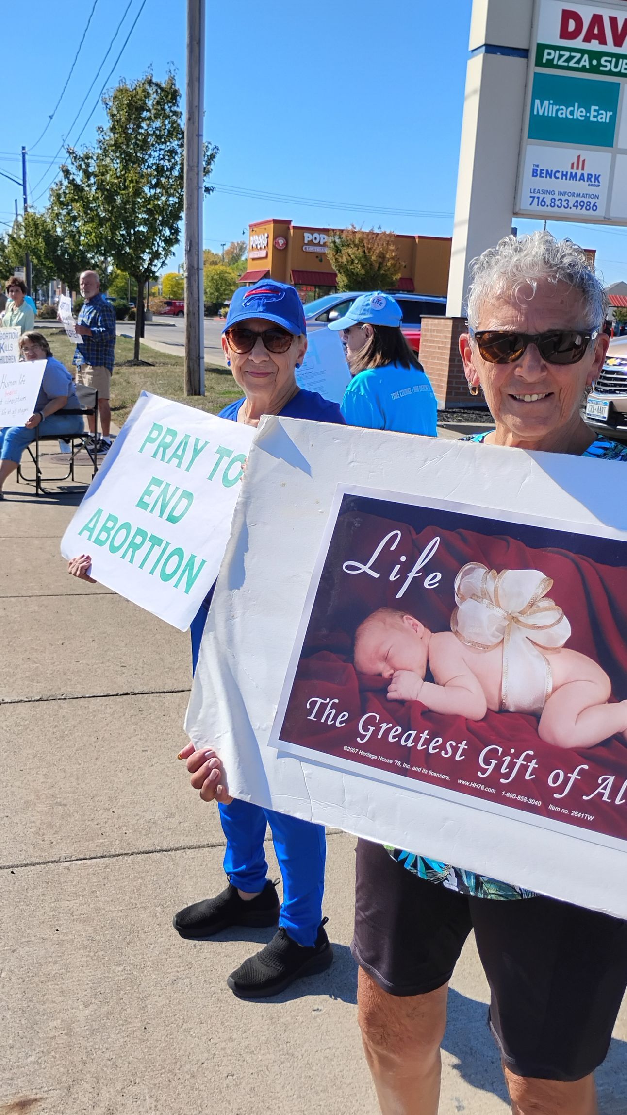Two people holding signs, protesting abortion, near a storefront on a sunny day. One sign says 