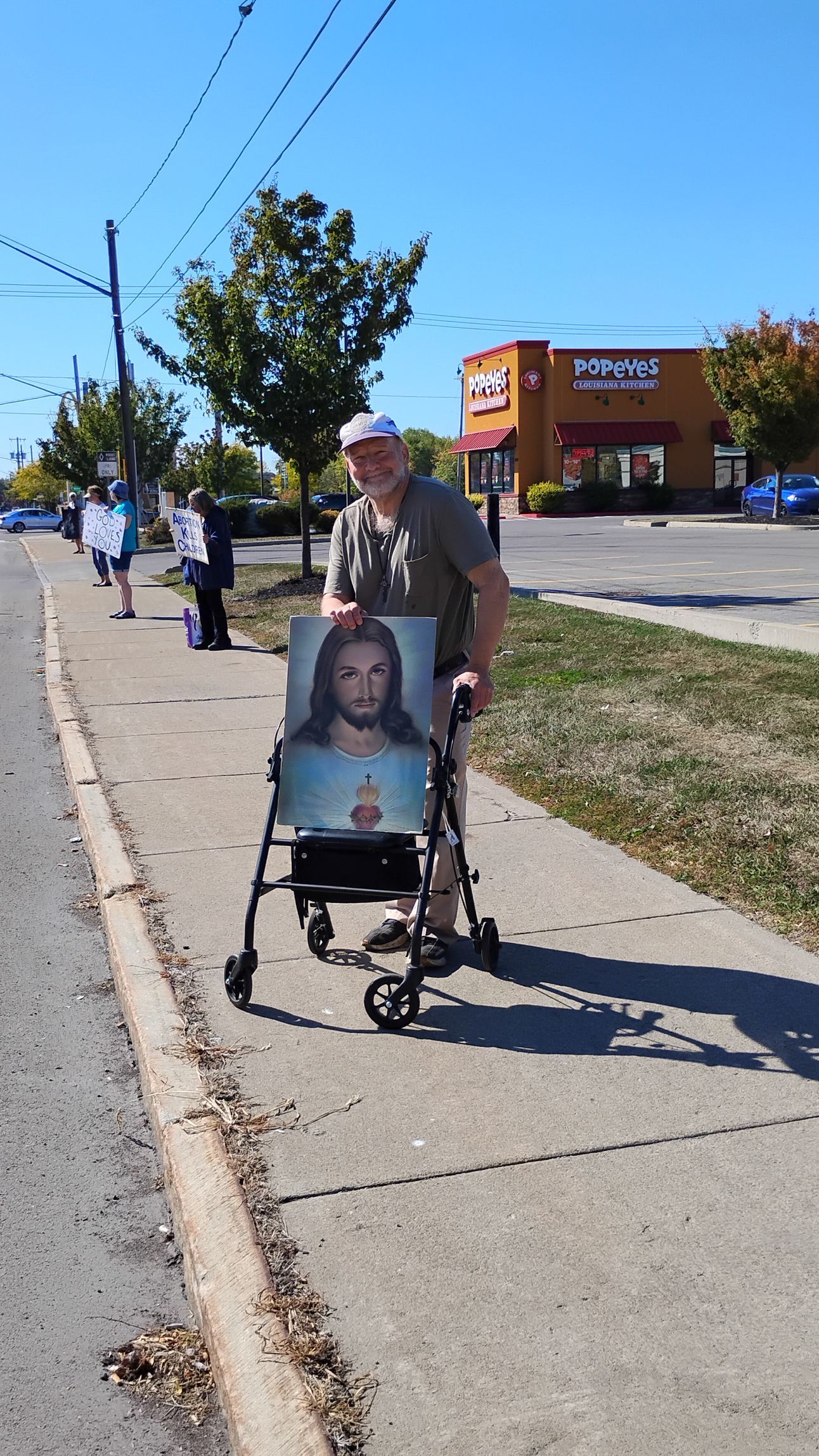 Man using a walker, holding a portrait, walking on a sidewalk. Sunny day, storefronts in the background.