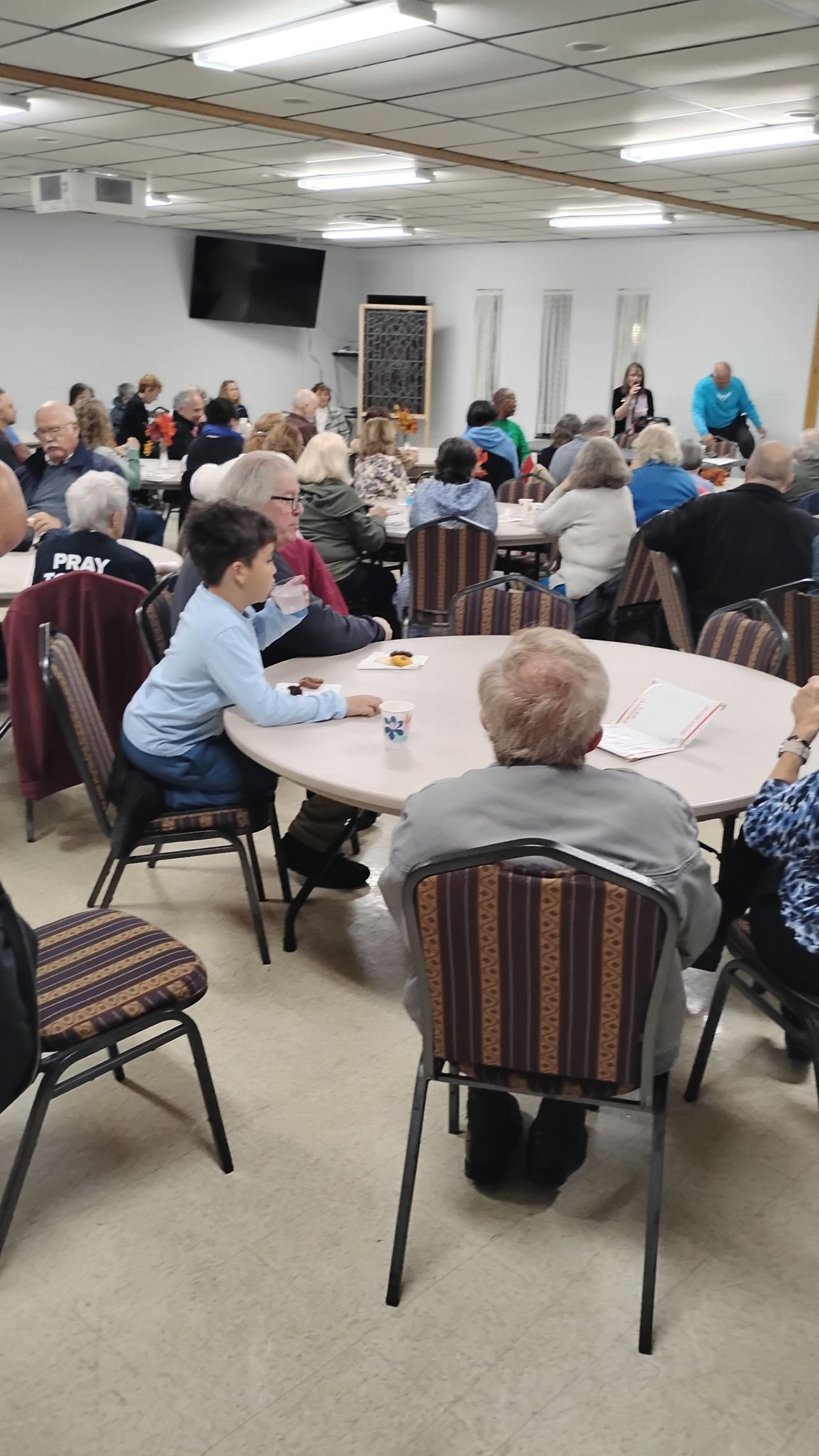 People seated at round tables in a large room, listening to a presentation.