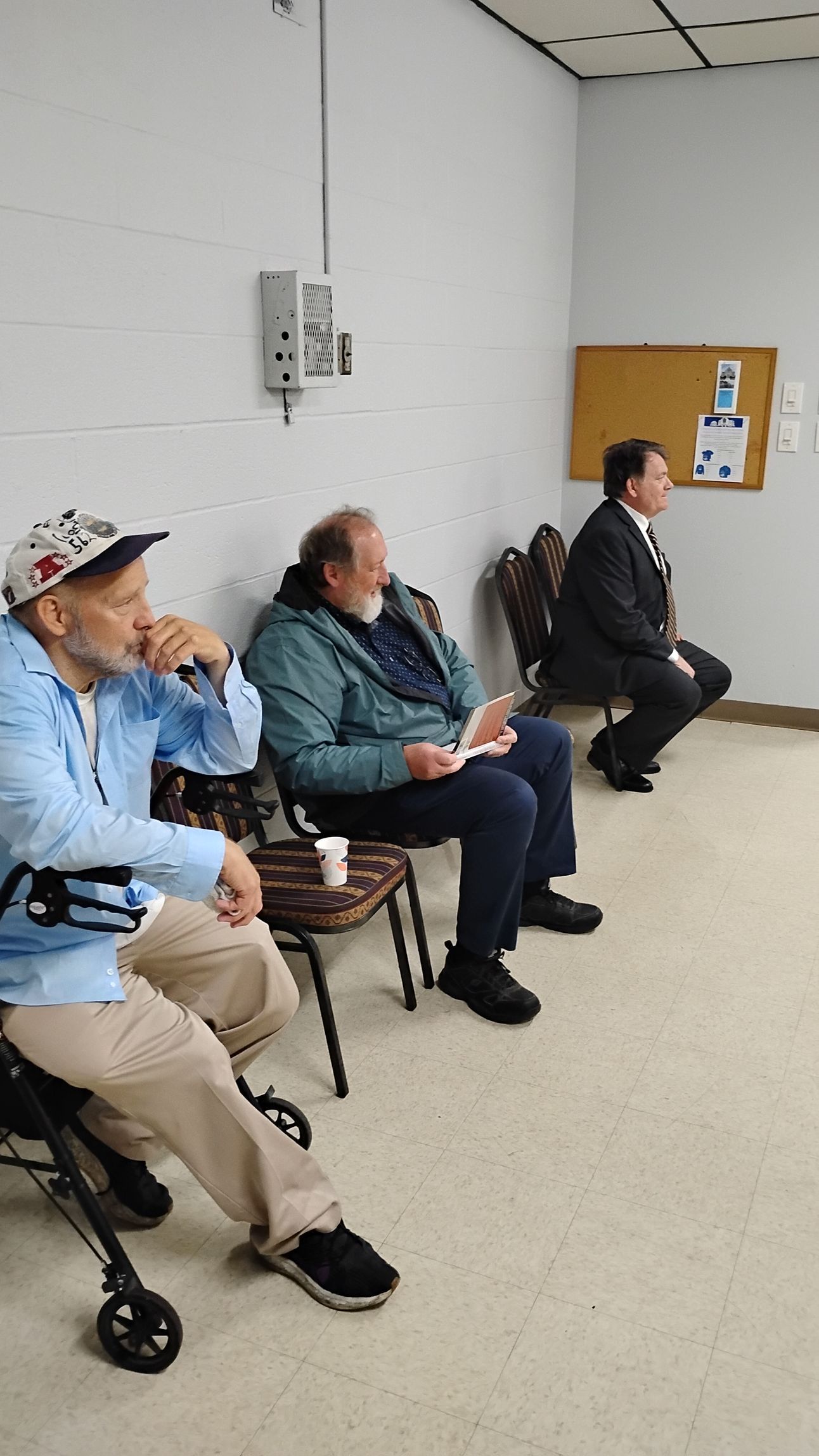 Three men seated indoors, likely at a meeting. One in a wheelchair, another with notes, and the third in a suit.