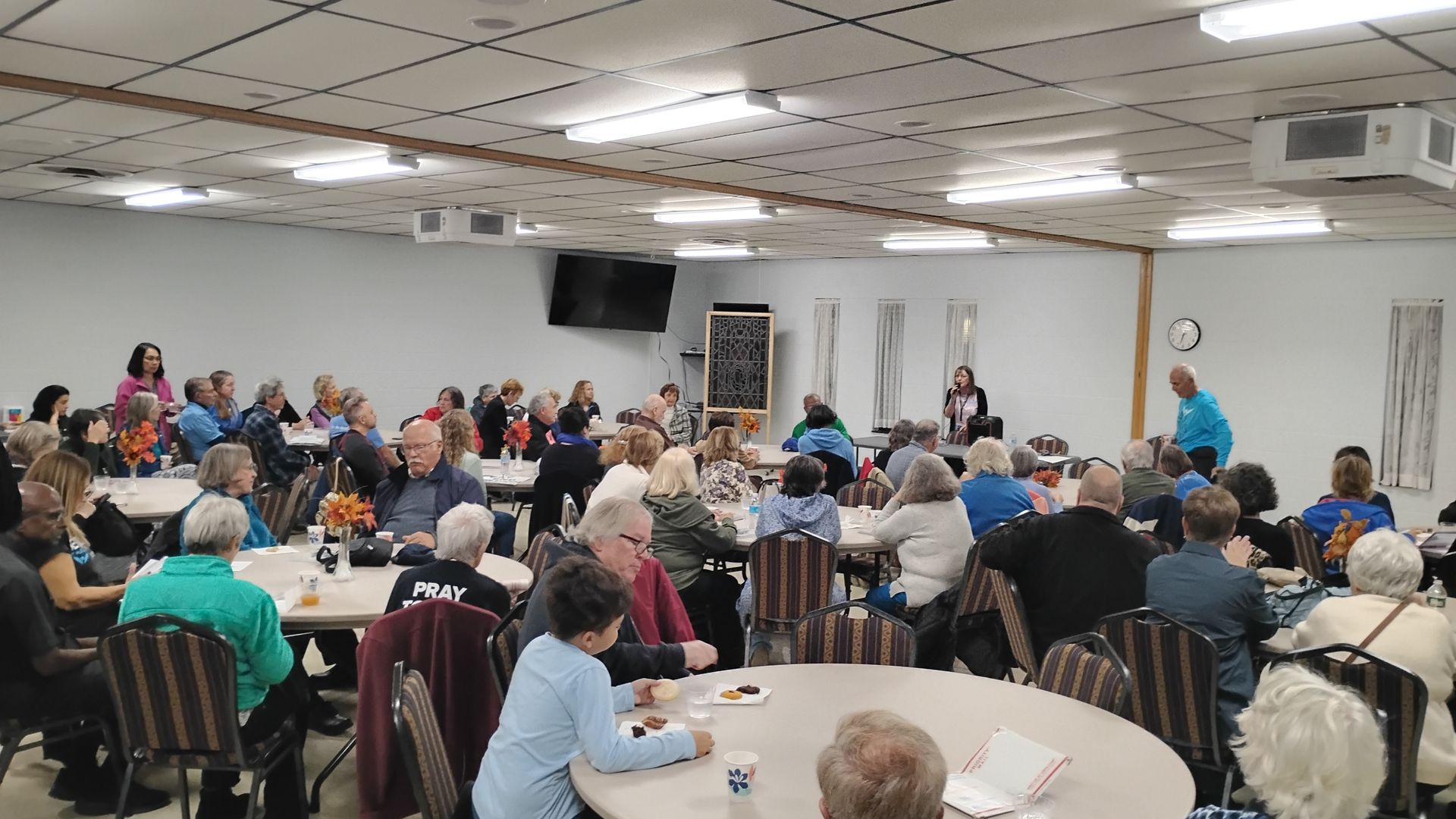 Large room with people seated at round tables. A woman speaks at a front table.