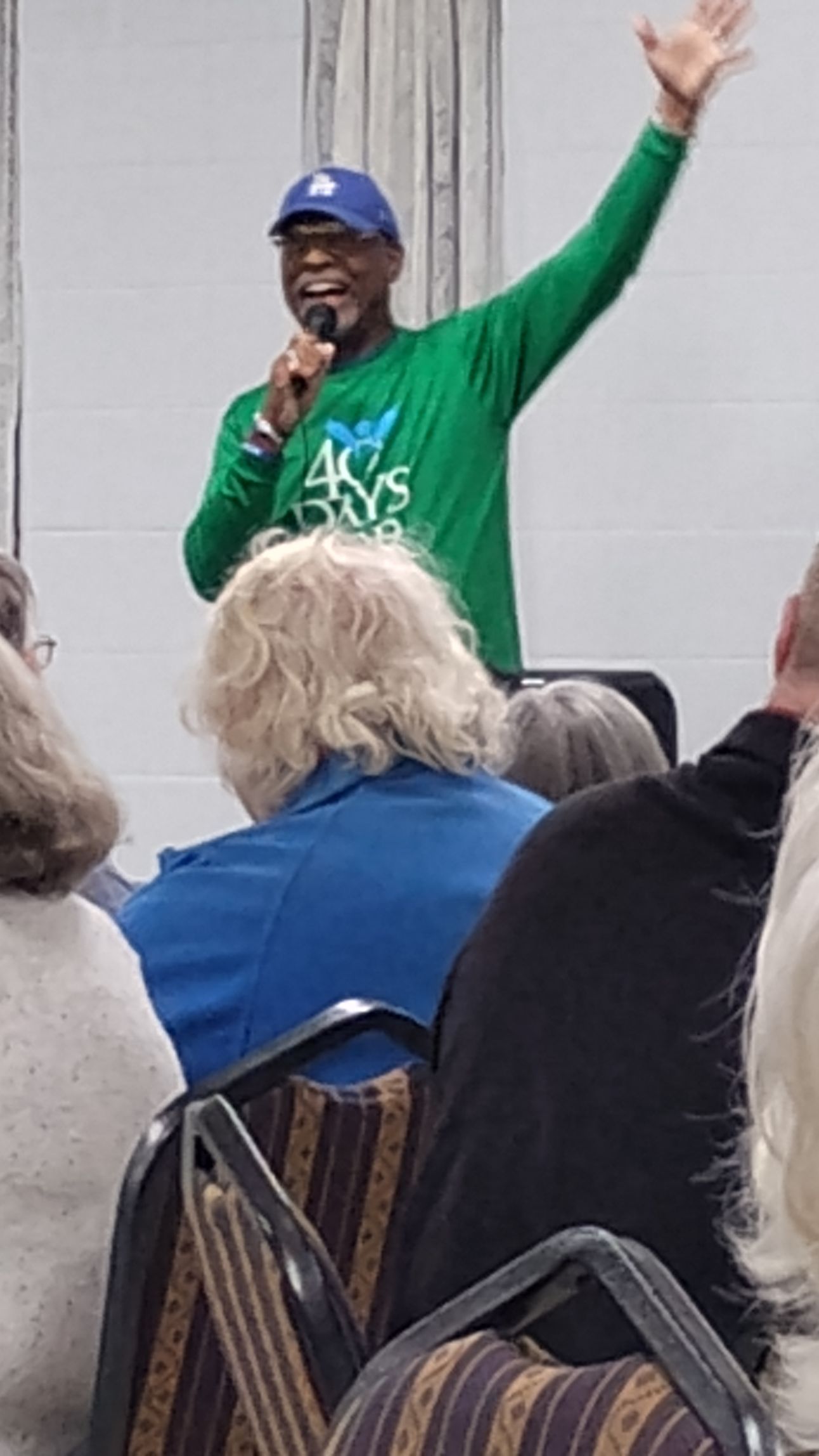 Man speaking at event, gesturing with arm raised. He's wearing a green shirt, blue cap, and holding a microphone.