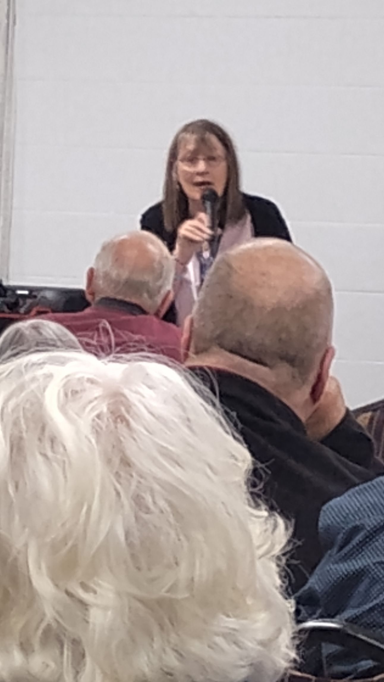 Woman speaking into a microphone, standing in front of a small audience.
