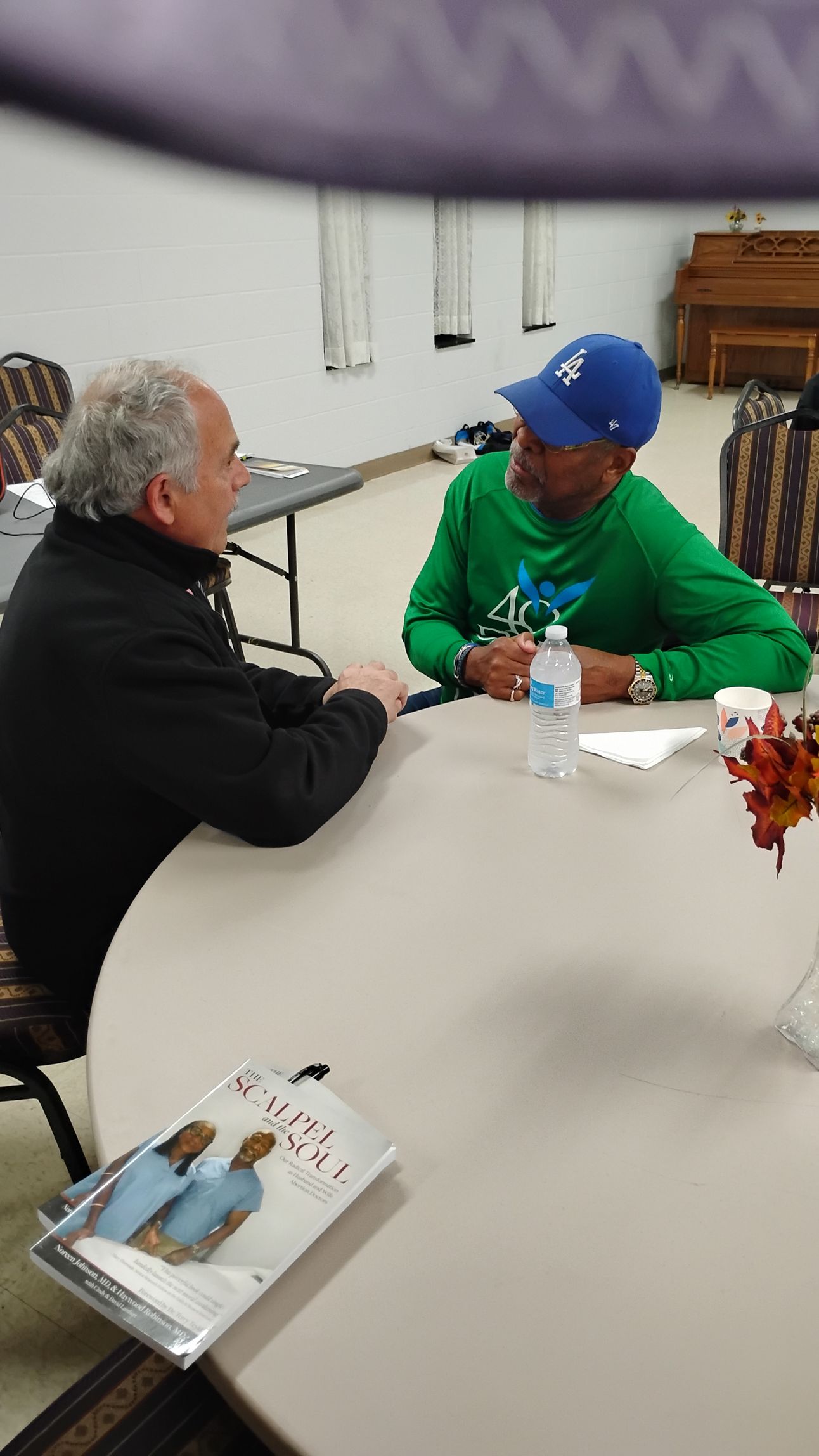 Two men seated at a round table, conversing. One wears a green shirt and blue cap, the other a black jacket. A book and water bottles are visible.