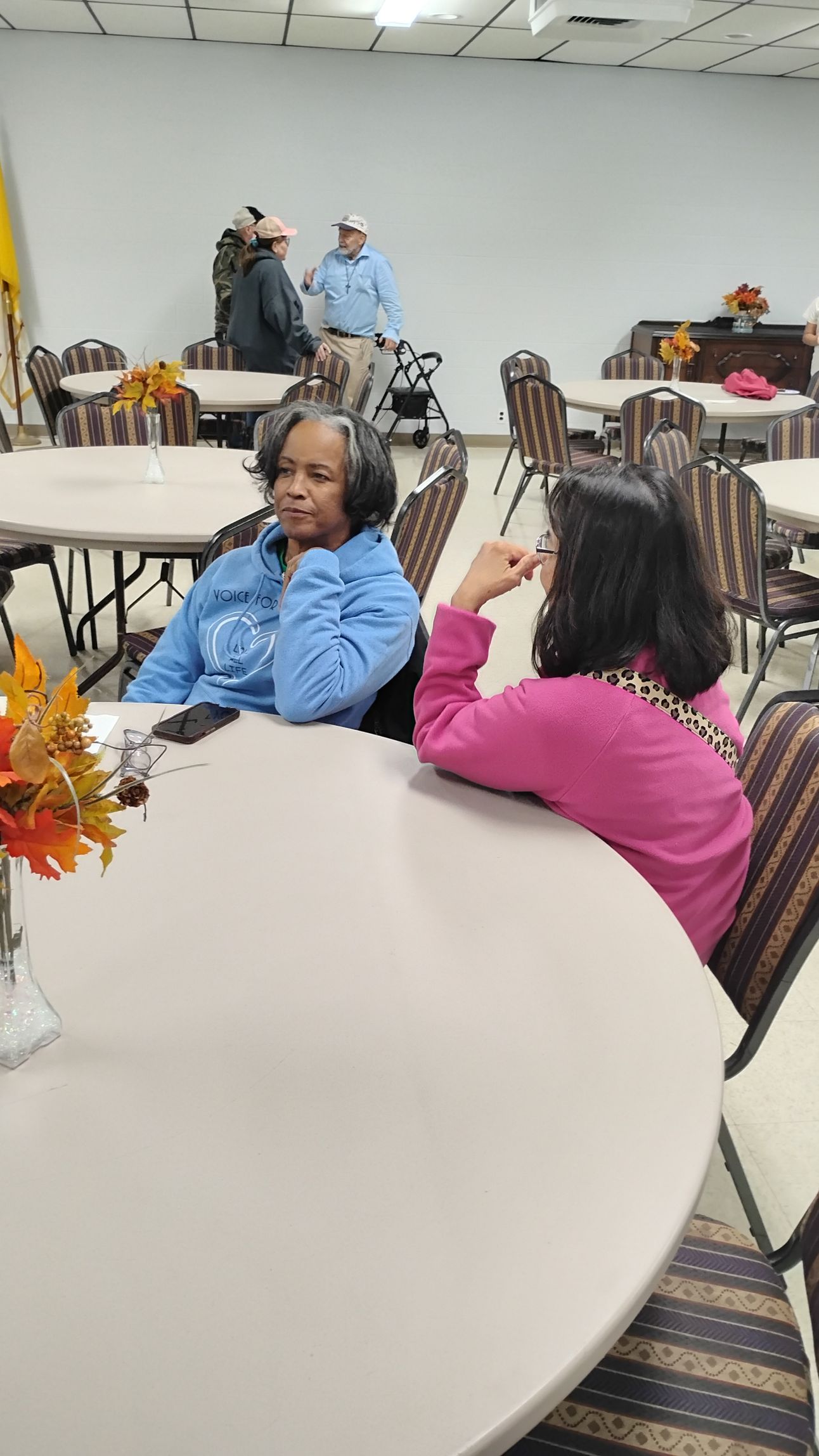 Two women seated at a table, conversing. Men in hats stand in the background. Beige tables, chairs, and white walls.