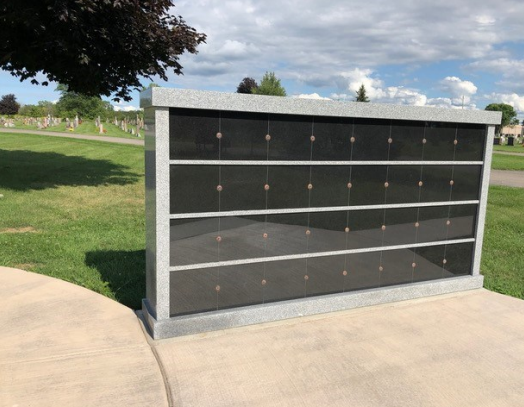 Exterior columbarium with black niches, set in a cemetery with green grass and a gray paved path.