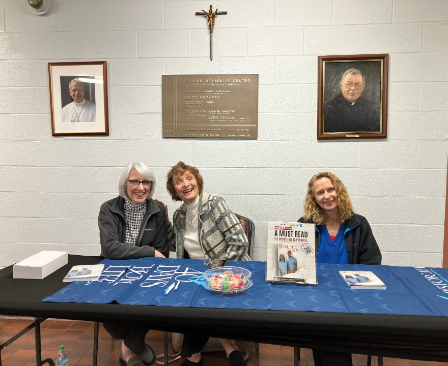 Three women smiling at a table with books and candy, under framed portraits and a crucifix.
