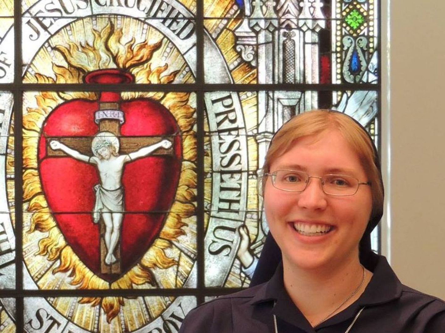 Nun smiles in front of a stained-glass window depicting the Sacred Heart of Jesus.