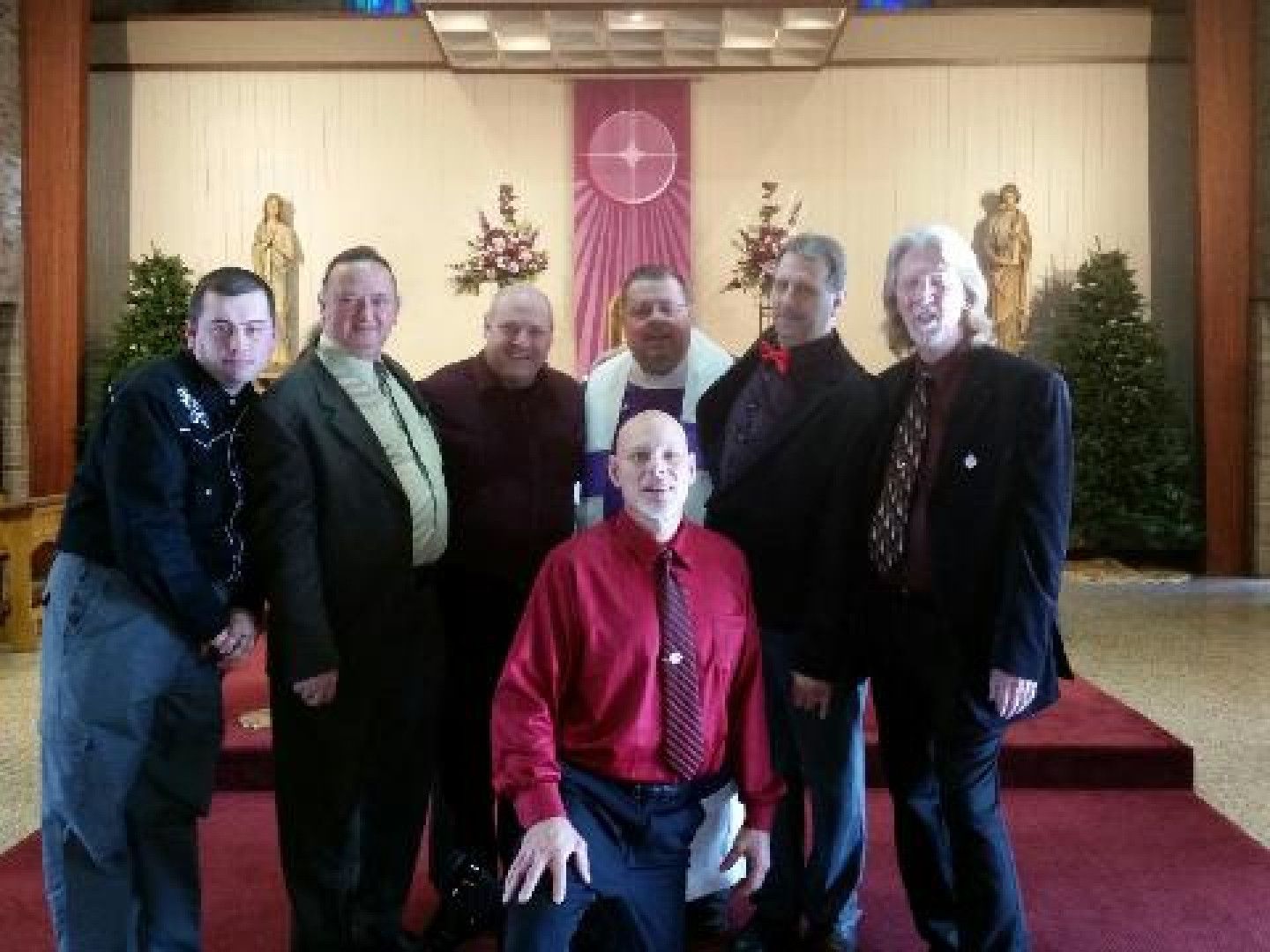 Group of men posing in a church; some wearing suits. One man in a wheelchair and one in clerical collar.
