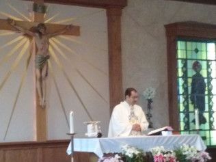 Priest at altar in church, crucifix on wall, stained glass window visible.