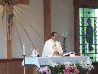 Priest at altar, crucifix, stained glass window. Flowers on table. White vestments, open book.