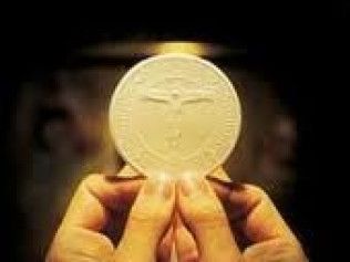 Hands holding a white Communion wafer with a cross imprint, illuminated against a dark background.