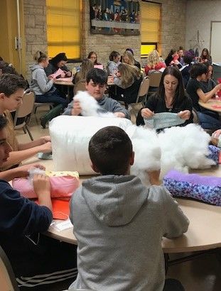 People stuffing pillows at tables in a room, with sunlight. Some focus on their work.