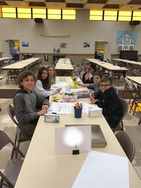 Students at a table, drawing. The room is a cafeteria with long tables and chairs. Smiling faces, art supplies.