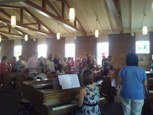 People singing in a church, a woman plays the piano. Light streams through windows.