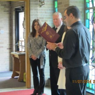 People in a church, ceremony: a man reads a document, others stand by.