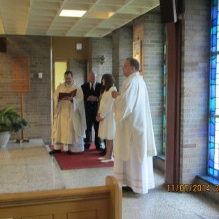 Clergy conduct a baptism in a church, with a person in white garments and a man in a black suit looking on.