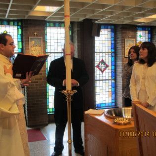 Clergy reading from a book during a baptism ceremony near a candle and stained glass windows.