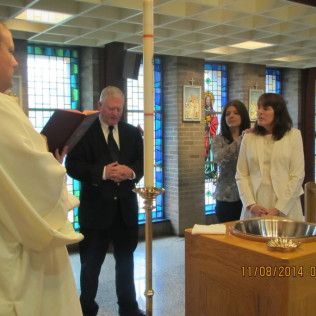 Baptism ceremony: Priest, man in suit, woman, and another woman stand near a baptismal font and candle in a church.
