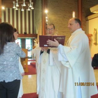 Clergy members in white vestments read from a book during a religious service. A woman stands nearby.