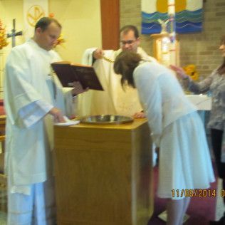 People at a baptism ceremony: A person in white is being baptized by a priest near a wooden stand with a basin.