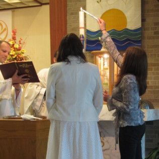 Ceremony in church: woman lighting candle for another woman standing by altar. Priest reading book.