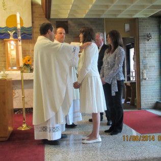 A person in white dress is being blessed by a priest. Ceremony inside church. Other people watch.